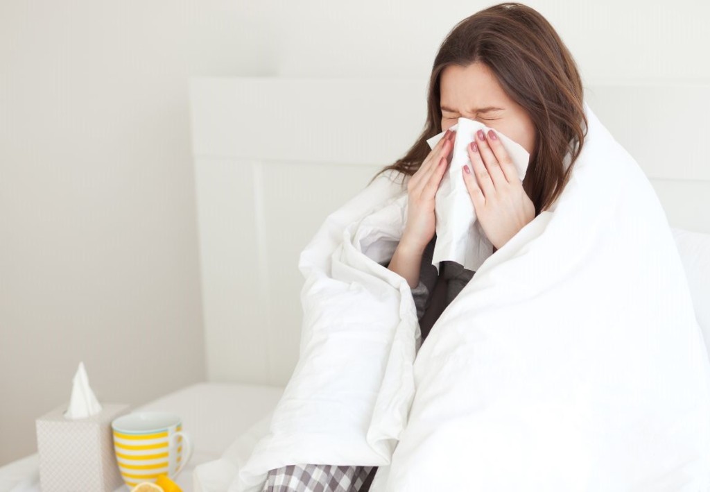 A woman sneezing into a tissue with a white blanket over her shoulders and hot lemon drink beside her